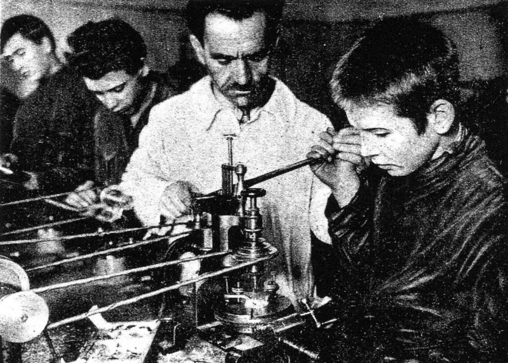 Transcription du magazine «Ogonyok» ci-dessus, 19 février 1928 : Il n'y a pas de production horlogère en URSS. Les montres de fabrication française et suisse sont très chères. Certaines entreprises coopératives achètent de vieilles montres, les "rafraîchissent" puis les revendent, également à un prix assez élevé. Nous devons créer notre propre industrie horlogère. À Moscou, ce travail a été lancé par un spécialiste de pointe de la technologie horlogère, V. O. Pruss, qui a fuit la Russie tsariste en tant que "hors-la-loi politique", et qui a 20 ans d'expérience dans les meilleures usines suisses. Le camarade Pruss enseigne la fabrication et l'assemblage de montres à des enfants sans abri dans l'atelier central du système scolaire MONO.  L'équipement et les matériaux pour l'atelier ont été apporté de Suisse par le camarade Pruss.